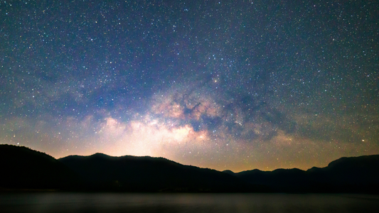 C'est un paysage montagneux , dans une nuit étoilée. Il y'a une lueur de lumière entre les montagnes. 